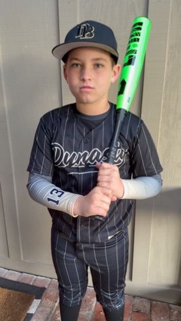 Young baseball player wearing a black pinstripe custom jersey with team name script text and the number 13 on the sleeve, holding a green bat.