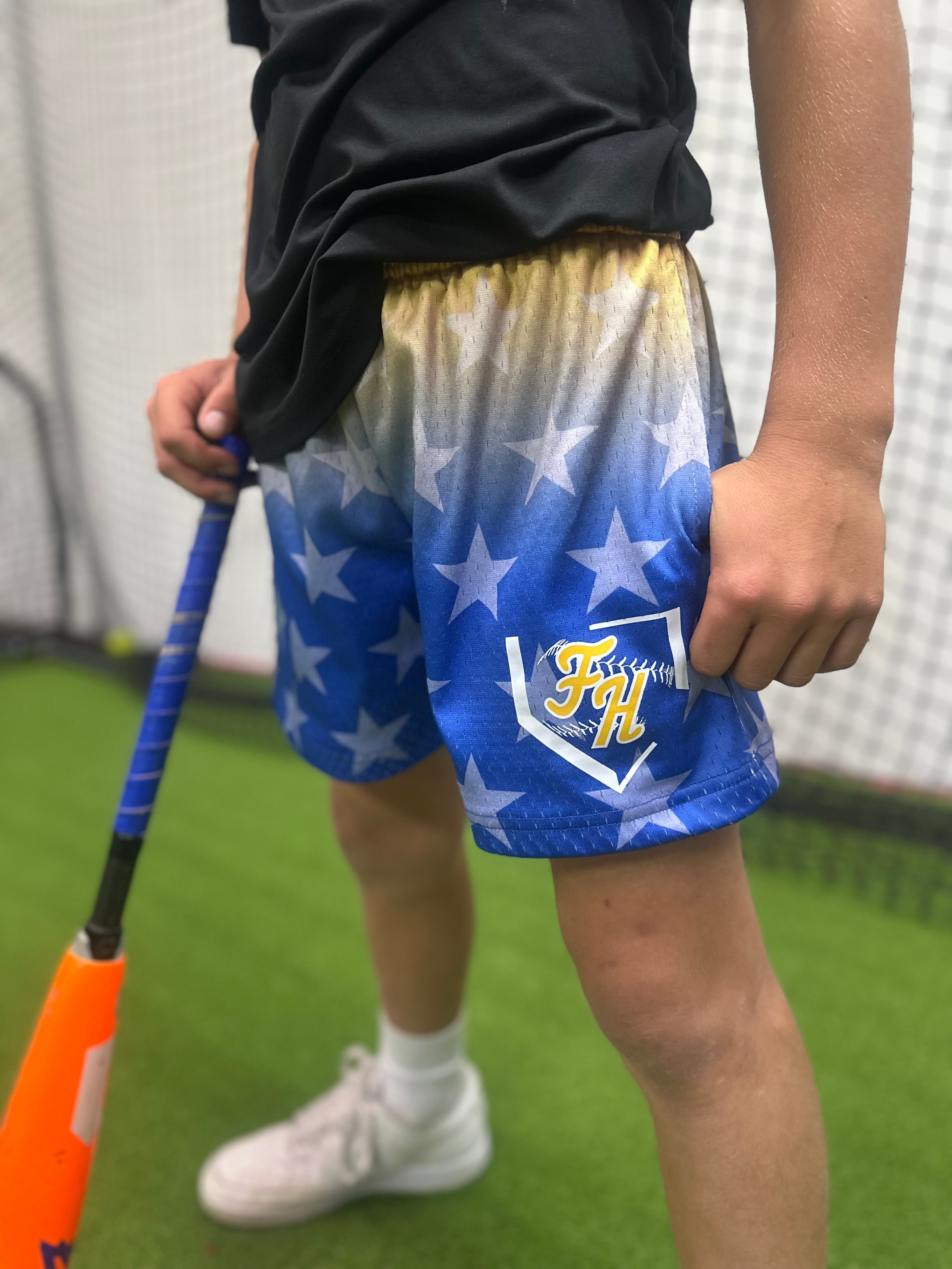 Person wearing blue shorts with white star pattern and logo, holding a baseball bat on a green field.