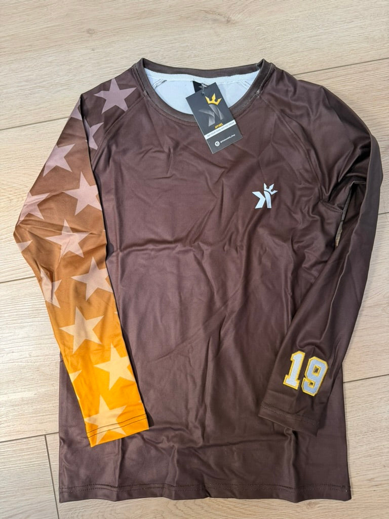 Brown long sleeve shirt with a gold star print ombre sleeve and the number 19 on the cuff, laid flat on a wood floor.
