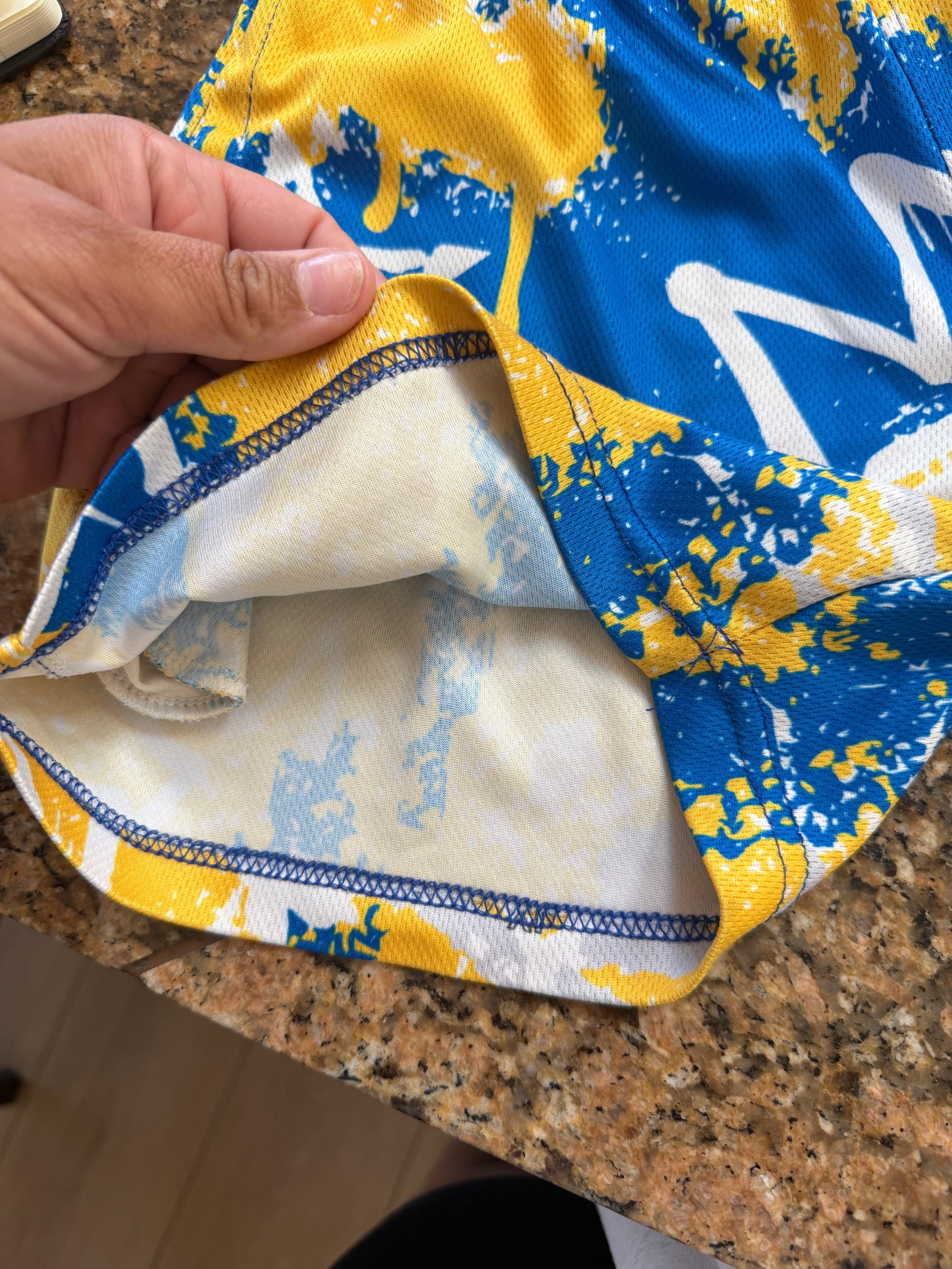 Close-up of a hand lifting the hem of blue and yellow abstract print mesh shorts to show the inner lining.