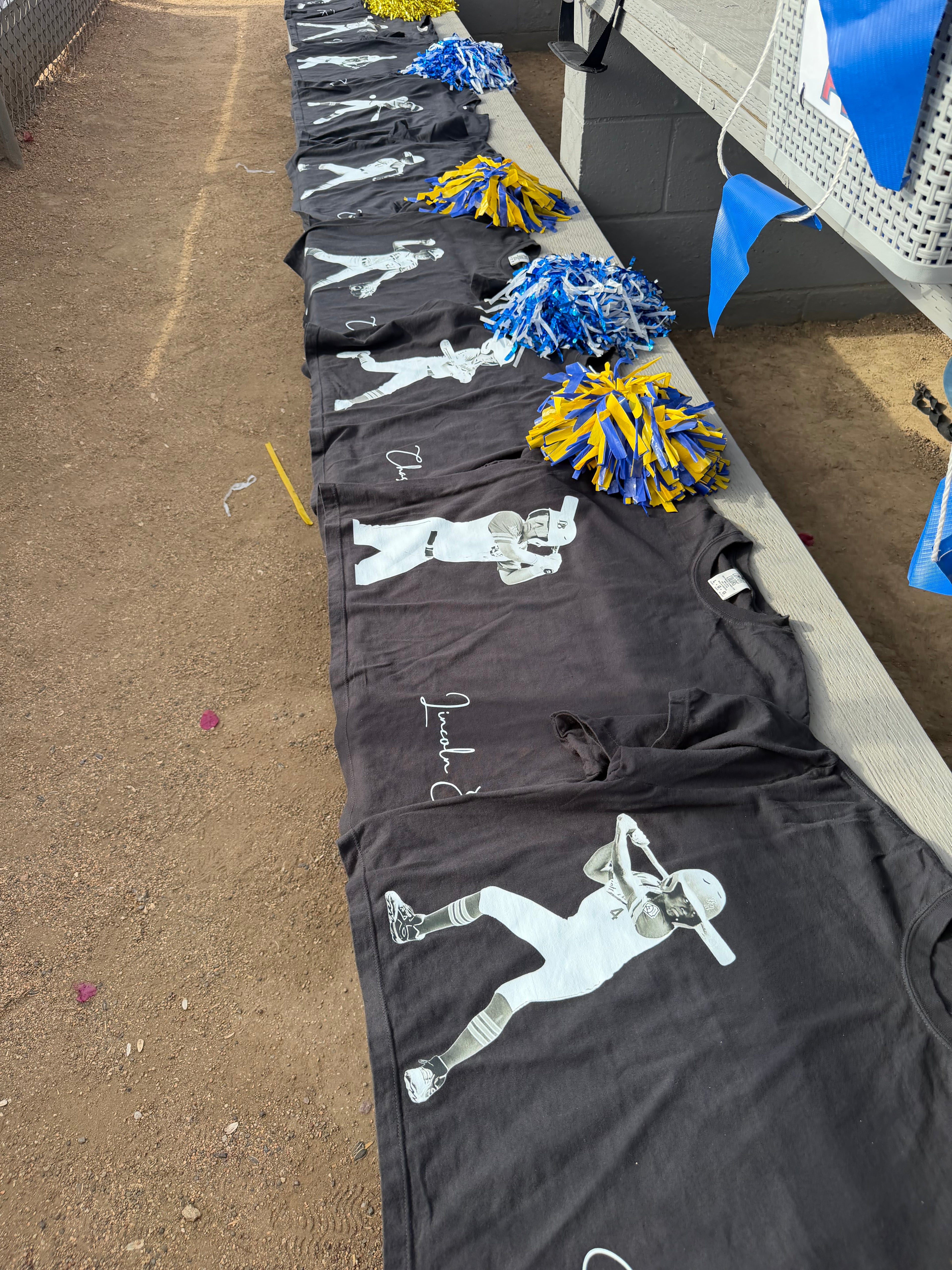 Row of black Signature Series tees laid out on bleachers, each printed with a white action-photo silhouette of a football player and a personalized signature.